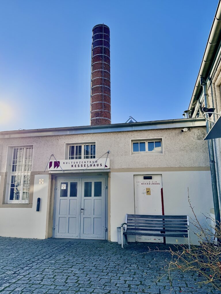 Brick chimney rises above a white cultural-center building under a clear blue sky. (Kulturzentrum Kesselhaus visible)