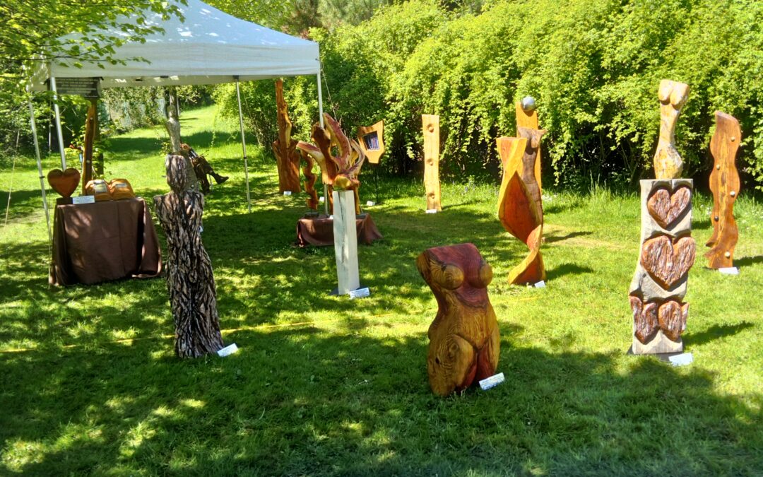 Outdoor art fair in a park with carved wooden sculptures arranged on the grass under a white canopy.