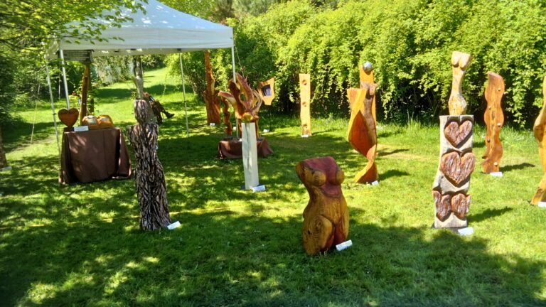 Outdoor art fair in a park with carved wooden sculptures arranged on the grass under a white canopy.