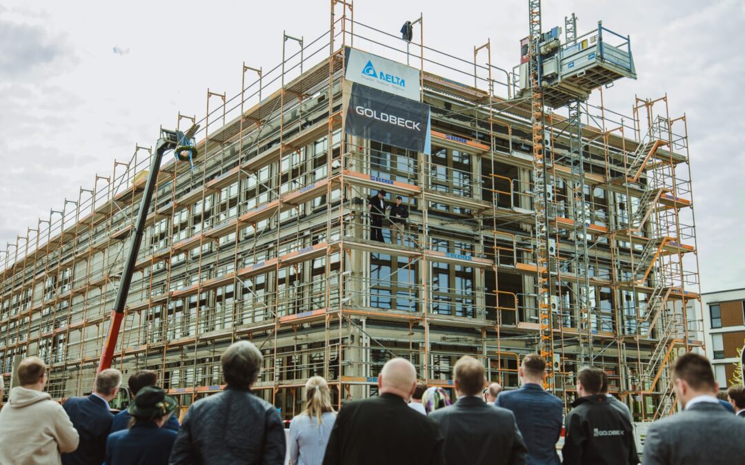 Construction site with multi-story scaffolding around a building; a crowd watches a ceremony or briefing on the ground level.