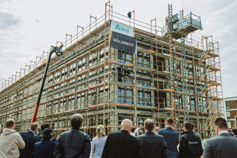 Construction site with multi-story scaffolding around a building; a crowd watches a ceremony or briefing on the ground level.