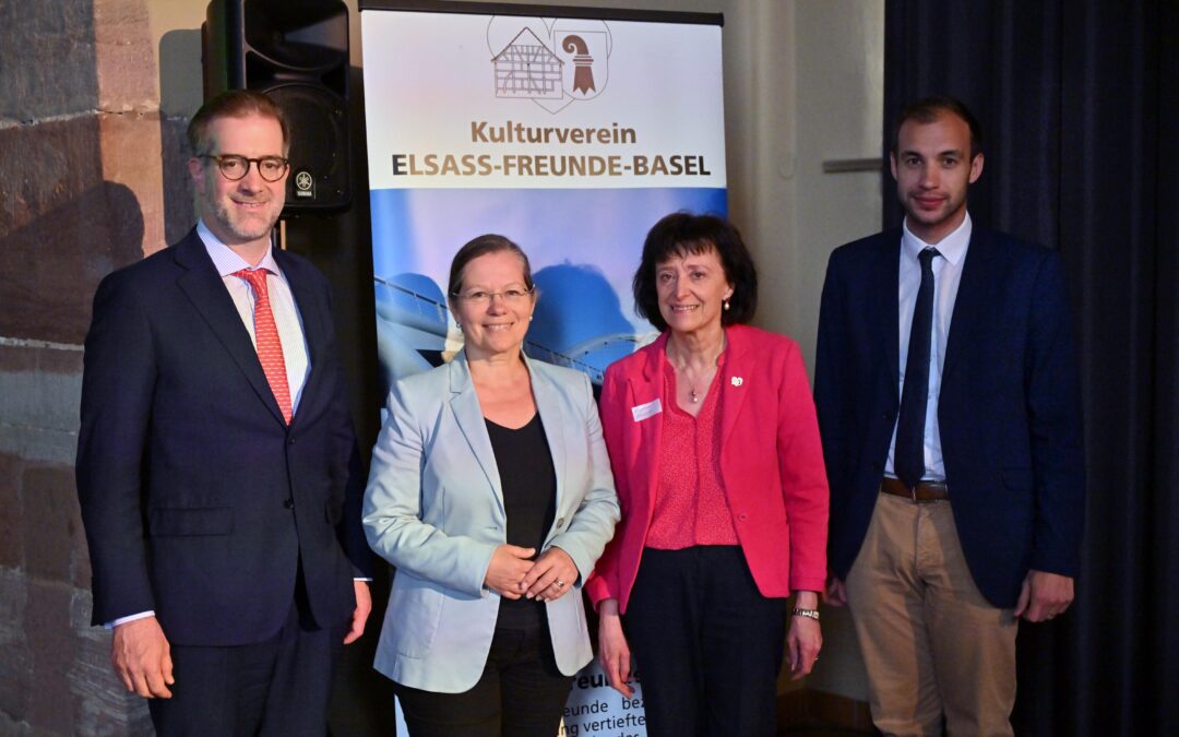 Group of four adults in business attire posing indoors in front of a banner that reads Kulturverein Elsass-Freunde-Basel.