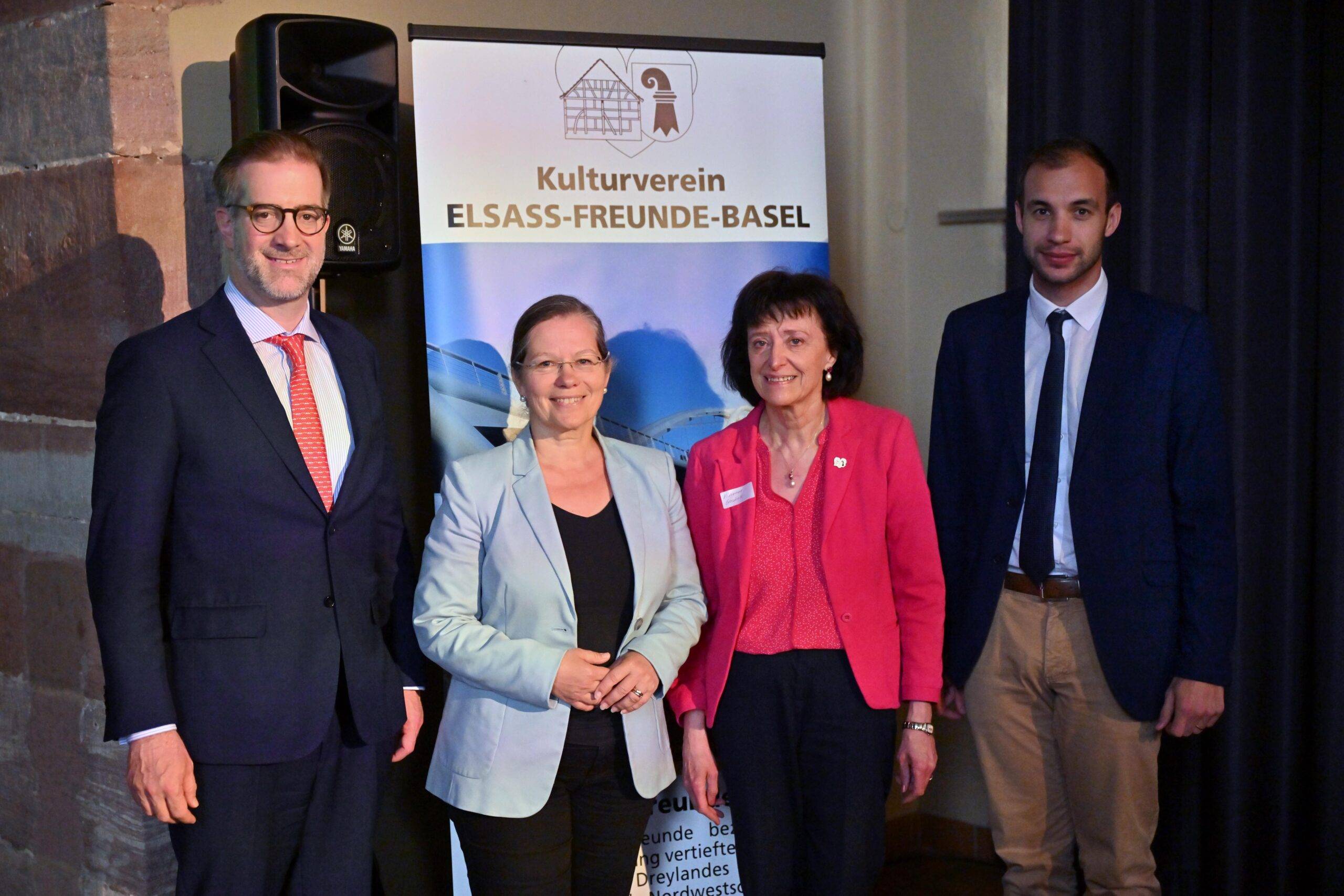 Group of four adults in business attire posing indoors in front of a banner that reads Kulturverein Elsass-Freunde-Basel.