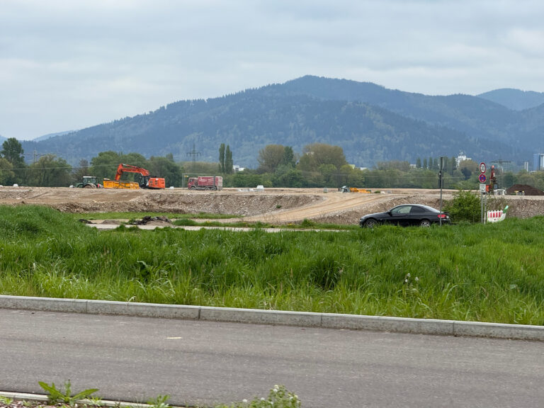 Construction site with orange excavator and truck on dirt lot, green grass in foreground and blue mountains in the distance under a cloudy sky.