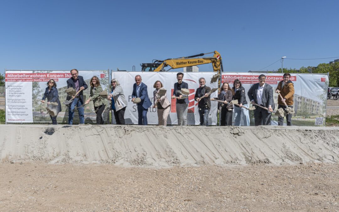 Group of people at a groundbreaking ceremony, each with a shovel, in front of construction banners and a yellow excavator under a clear blue sky.