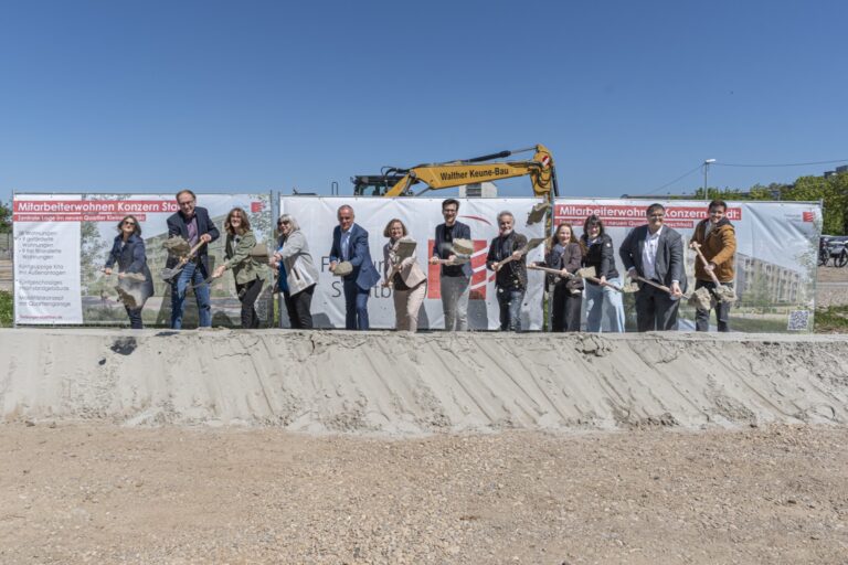 Group of people at a groundbreaking ceremony, each with a shovel, in front of construction banners and a yellow excavator under a clear blue sky.