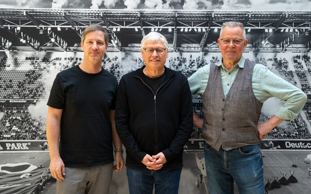 Three men stand side by side in front of a large black-and-white stadium backdrop/photo mural.
