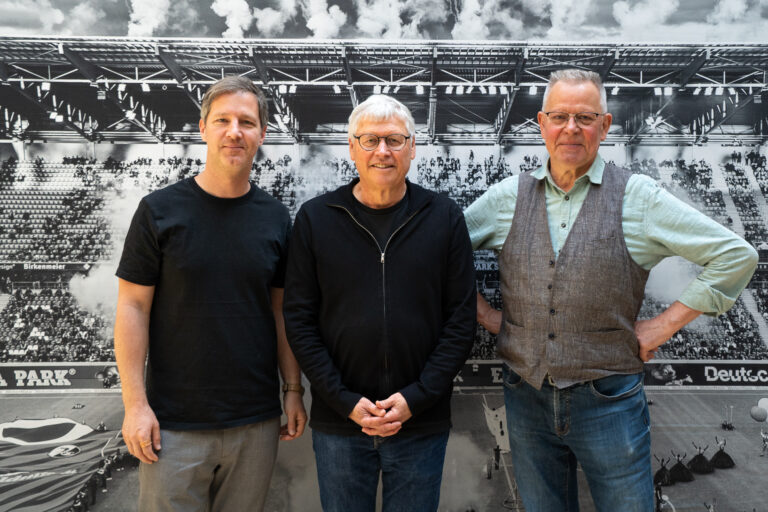 Three men stand side by side in front of a large black-and-white stadium backdrop/photo mural.