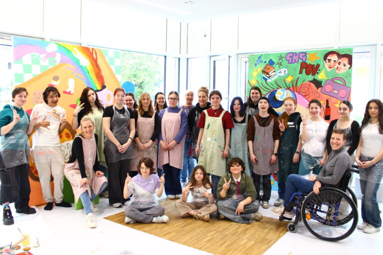 Group of students in colorful aprons posing for a photo in an art studio with bright mural paintings. Wheelchair visible on the right edge.