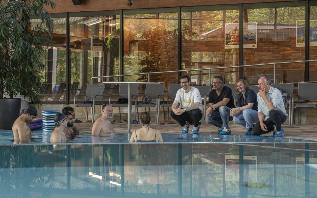 Poolside pool therapy class: adults sit on edge and chat while swimmer-participants stand in the water by the lane, indoor facility with plants and brick walls