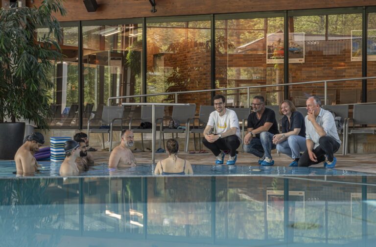 Poolside pool therapy class: adults sit on edge and chat while swimmer-participants stand in the water by the lane, indoor facility with plants and brick walls