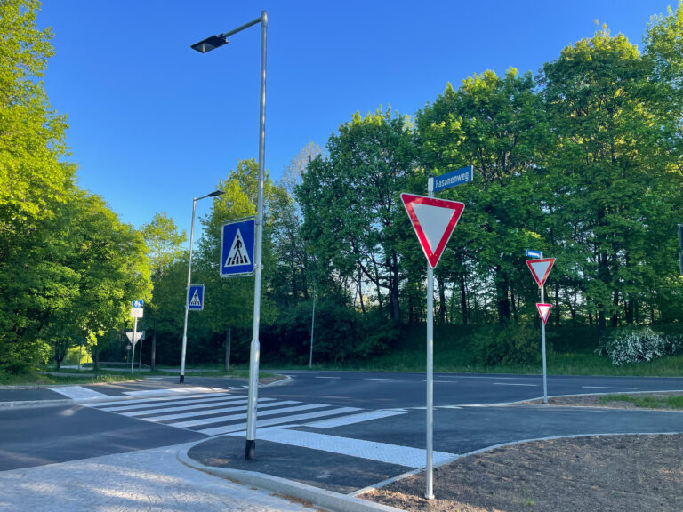 Sunny intersection with yield sign and pedestrian crossing signs beside a tree-lined street labeled Fasanenweg in bright daylight.