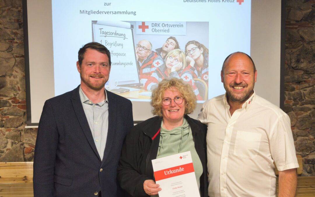 Four adults smiling for a photo at a DRK event; the woman in front holds a white certificate labeled 'Urkunde' with red cross logo visible.