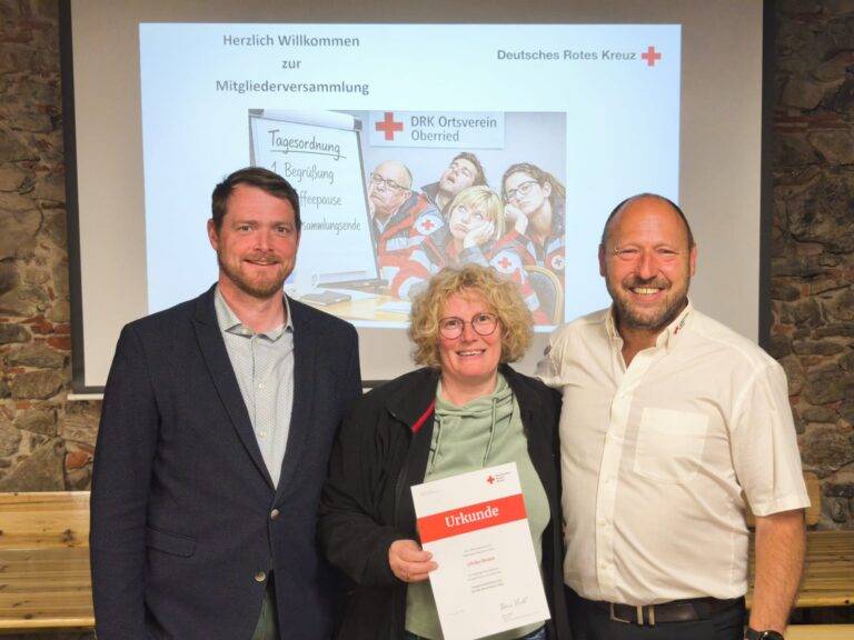 Four adults smiling for a photo at a DRK event; the woman in front holds a white certificate labeled 'Urkunde' with red cross logo visible.