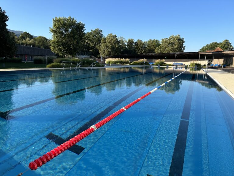 Outdoor swimming pool with red lane ropes, calm blue water, and surrounding trees on a sunny day.