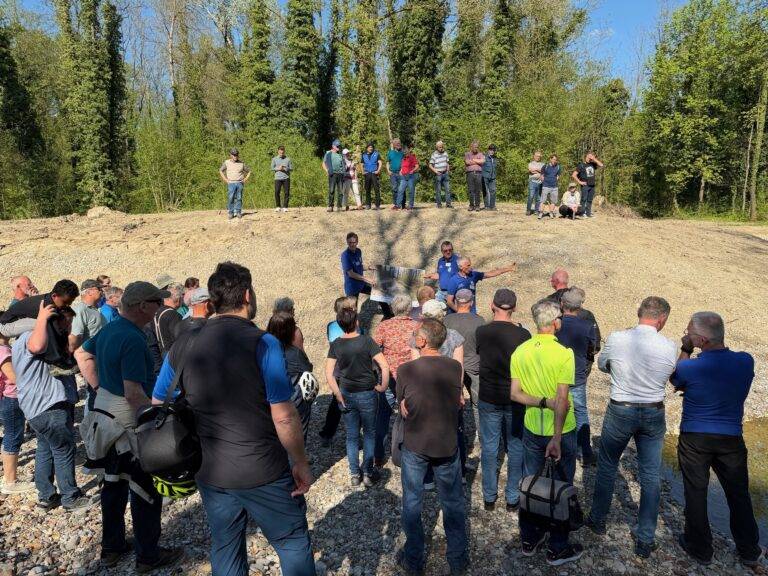 Group of people gathered on a dirt mound listening to two presenters in blue shirts near a display board, trees in the background.