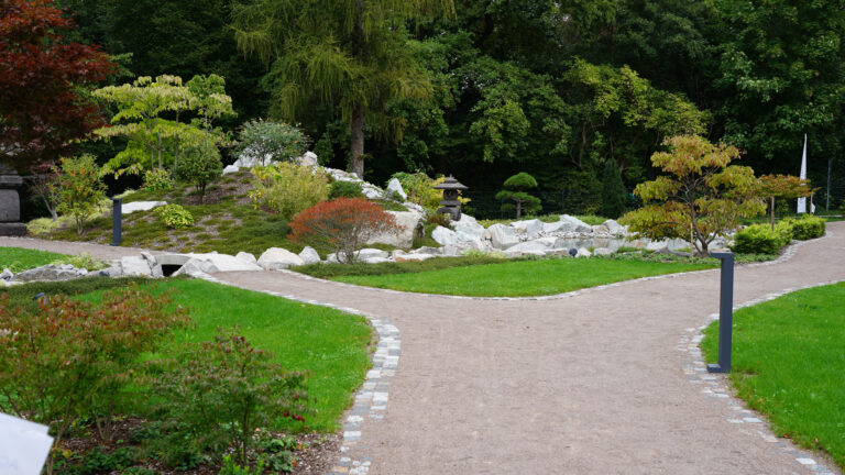 Curved gravel garden path lined with stones, green lawns, and a Japanese-style rock lantern among trees.