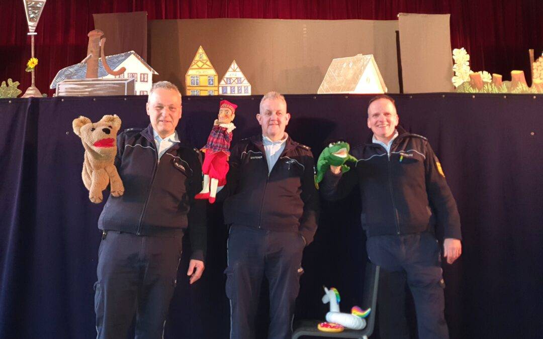 Four men in matching jackets pose on a stage, each holding a hand puppet in front of a blue curtain set with cardboard houses and stage props in the background.