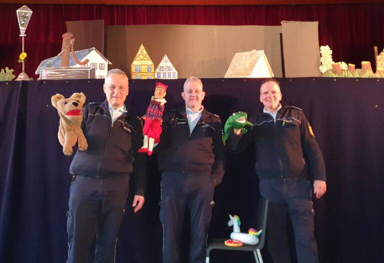 Four men in matching jackets pose on a stage, each holding a hand puppet in front of a blue curtain set with cardboard houses and stage props in the background.