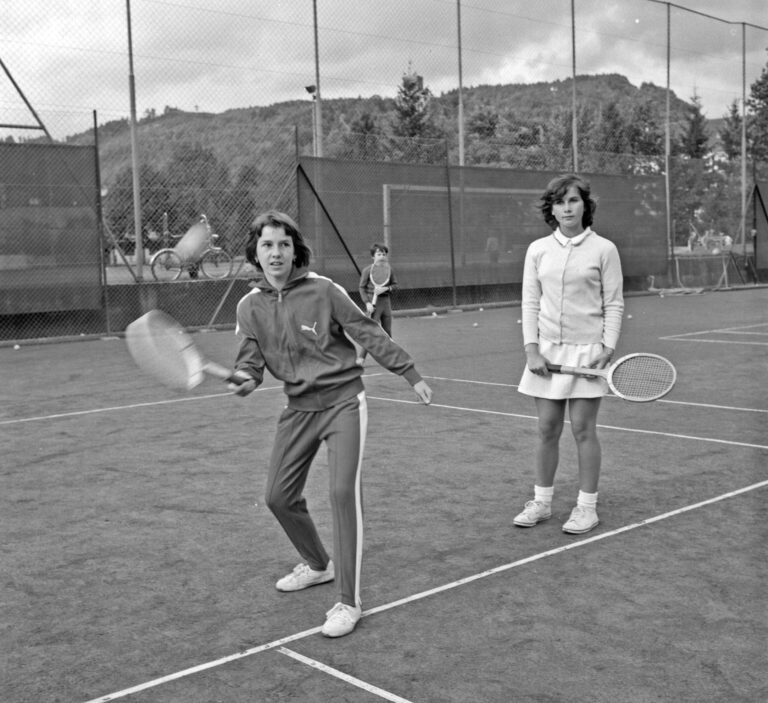Two women stand on a tennis court with rackets, near a chain-link fence, hills in the background (black-and-white).