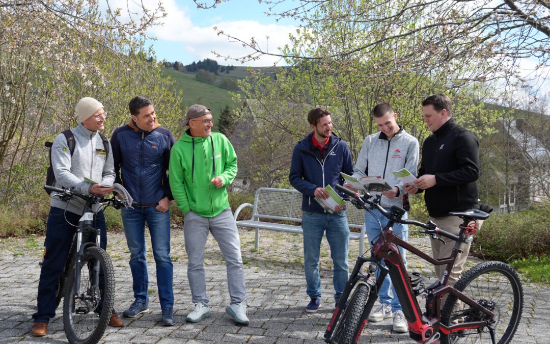 Six men with bicycles stand together outdoors, smiling while reading brochures on a sunny hillside path.