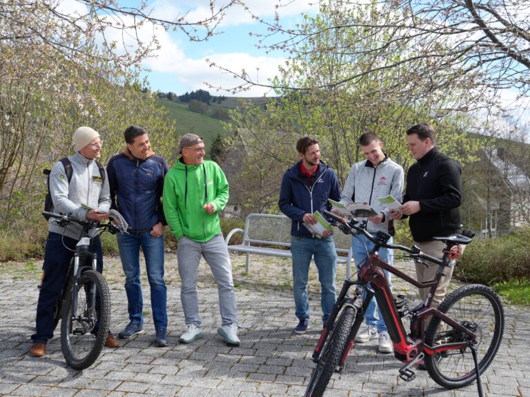 Six men with bicycles stand together outdoors, smiling while reading brochures on a sunny hillside path.