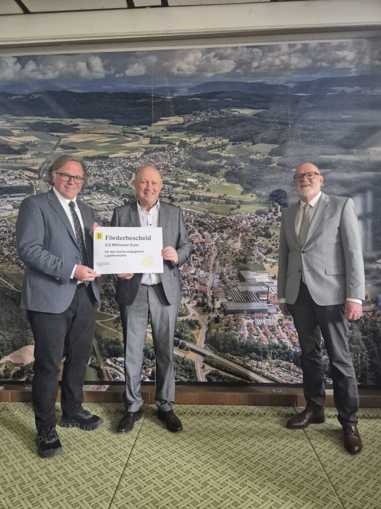 Three men in suits pose indoors in front of a large aerial-mural backdrop, with the middle man holding a certificate that reads 'Förderbescheid' for 3.5 million euros.