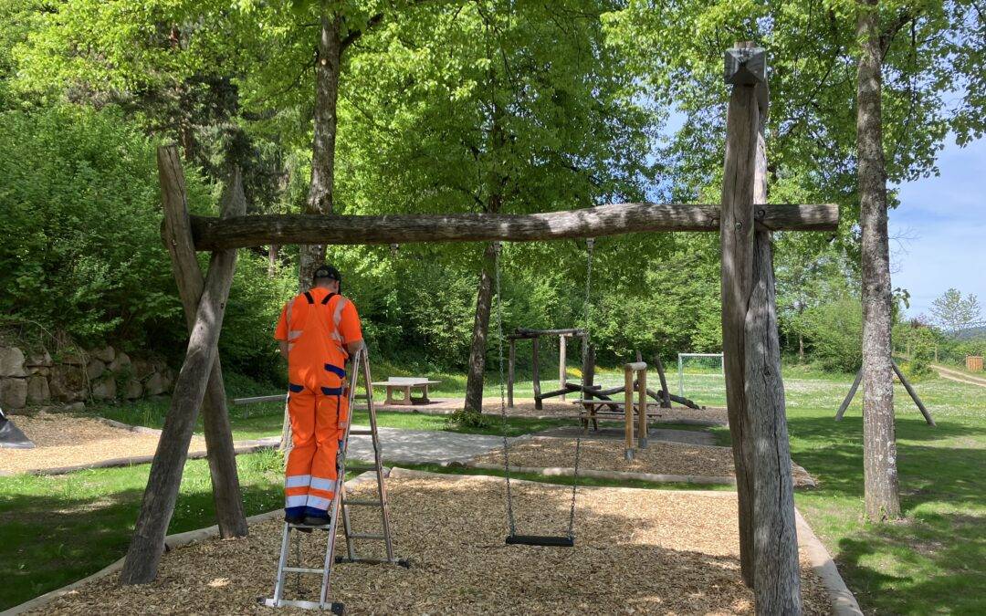 Worker in bright orange safety gear stands on a ladder beside a wooden swing set in a park area with trees above.