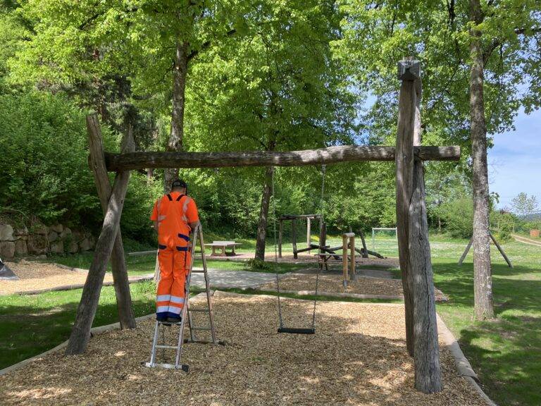 Worker in bright orange safety gear stands on a ladder beside a wooden swing set in a park area with trees above.