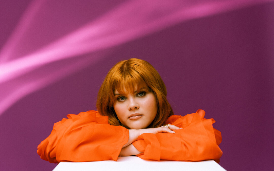 Person with orange-red hair rests chin on folded arms on a white surface, wearing bright orange sleeves against a purple backdrop.