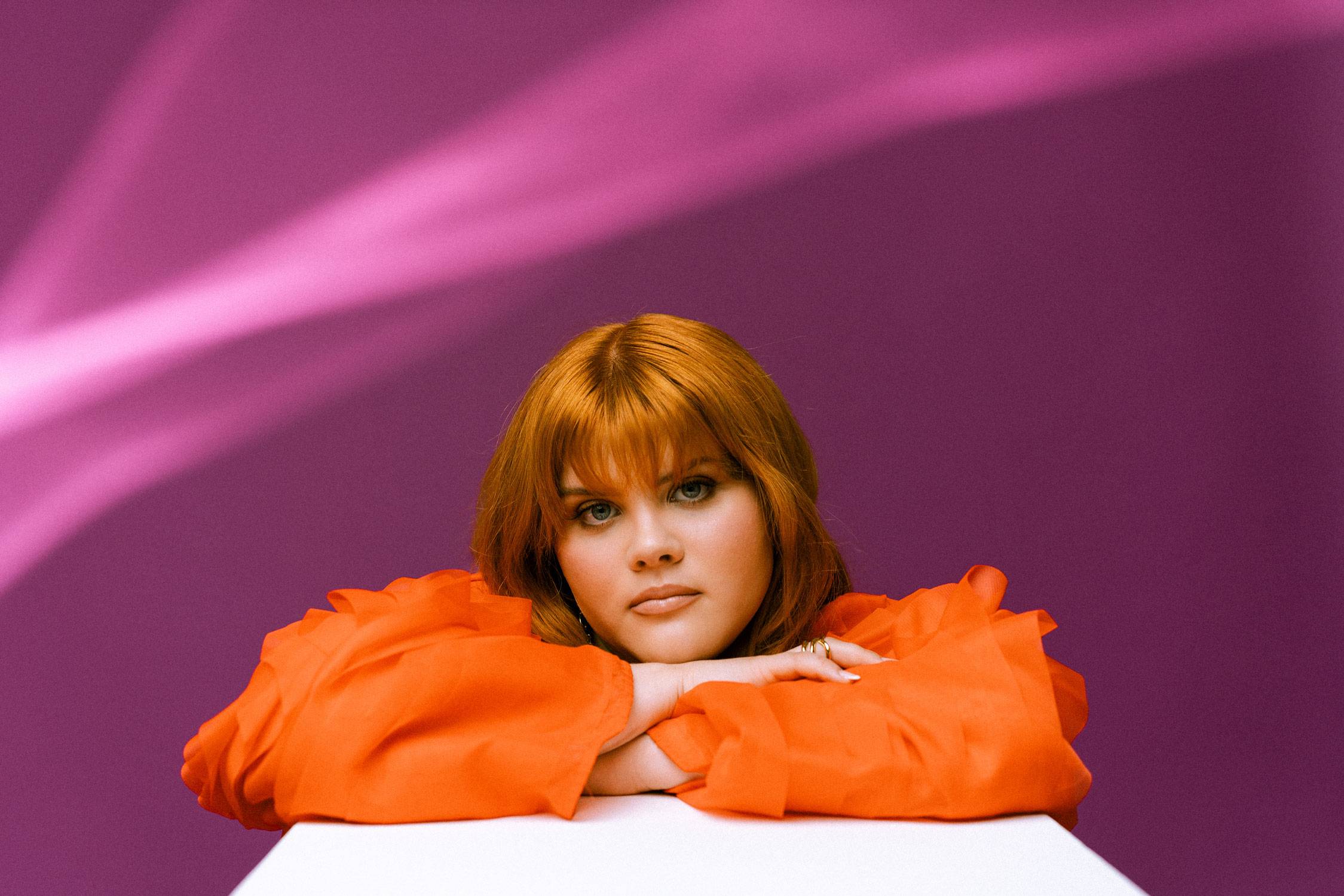 Person with orange-red hair rests chin on folded arms on a white surface, wearing bright orange sleeves against a purple backdrop.