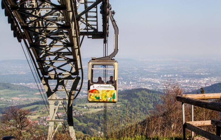 Cable car suspended from a tall steel tower over a valley with a distant town below and fields stretching out beneath.