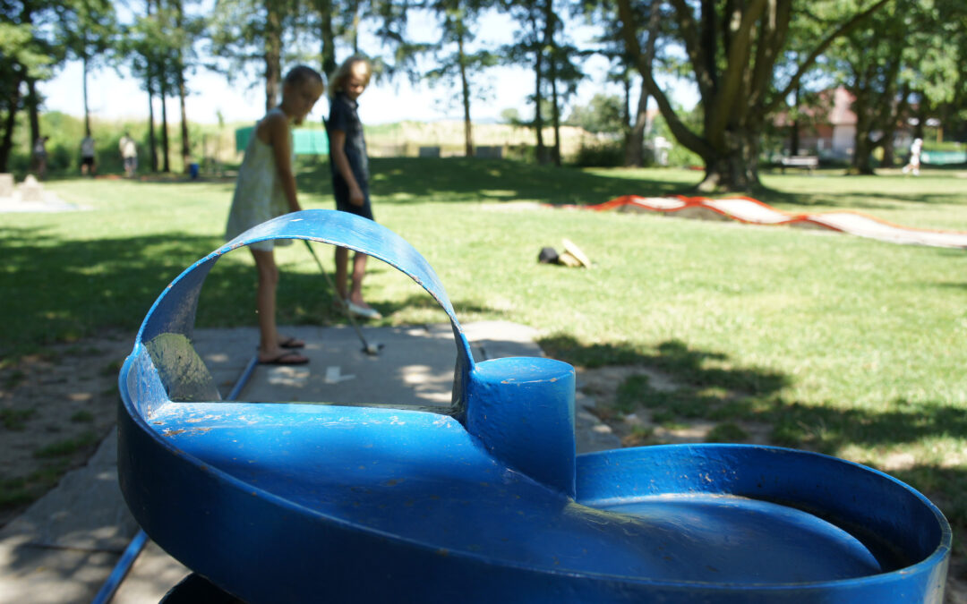 Close-up of a blue circular playground ride in the foreground with a grassy park and blurred kids in the background (sunny day).