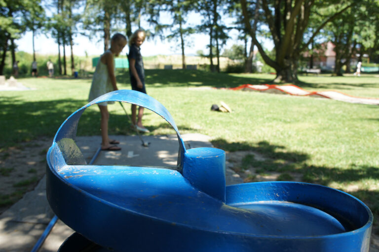 Close-up of a blue circular playground ride in the foreground with a grassy park and blurred kids in the background (sunny day).