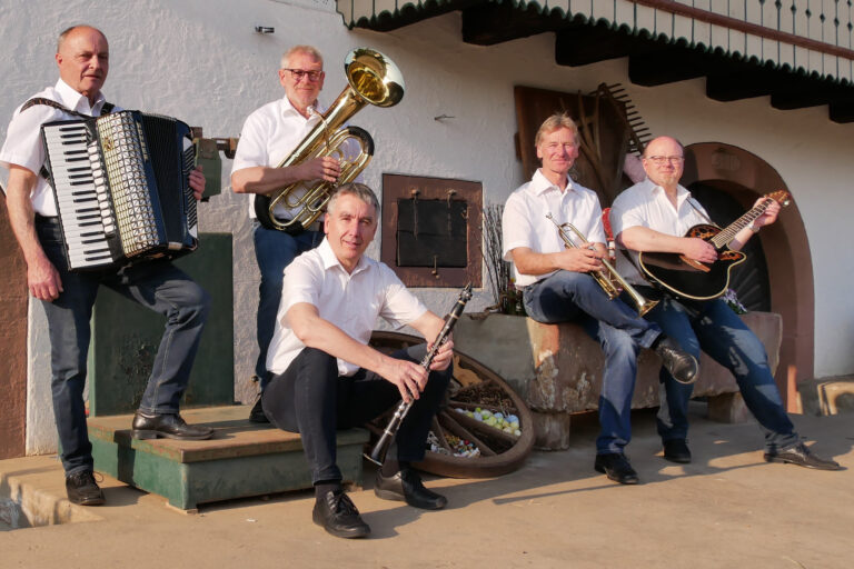 Five male musicians in white shirts pose with accordion, tuba, trumpet, clarinet and guitar outside a white-washed building.