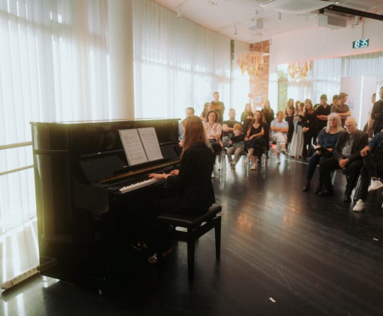 Pianist seated at a black grand piano, reading sheet music, while a seated and standing audience watches in a bright, modern room.