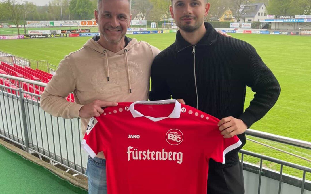 Two men posing on a stadium balcony, holding a red soccer jersey with 'Fürstenberg' sponsor logo.