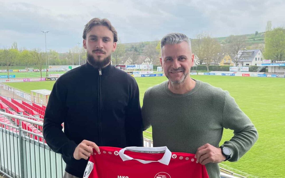 Two men stand on a stadium balcony holding a red soccer jersey with white accents and the word 'Fürstenberg' visible.