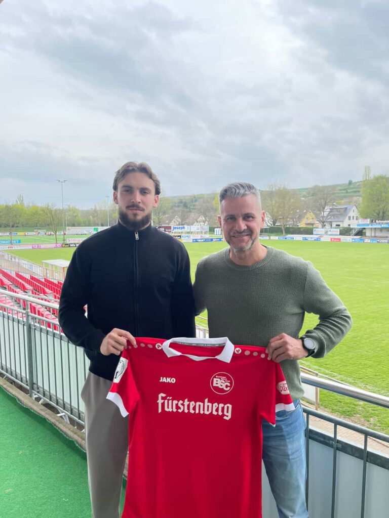 Two men stand on a stadium balcony holding a red soccer jersey with white accents and the word 'Fürstenberg' visible.