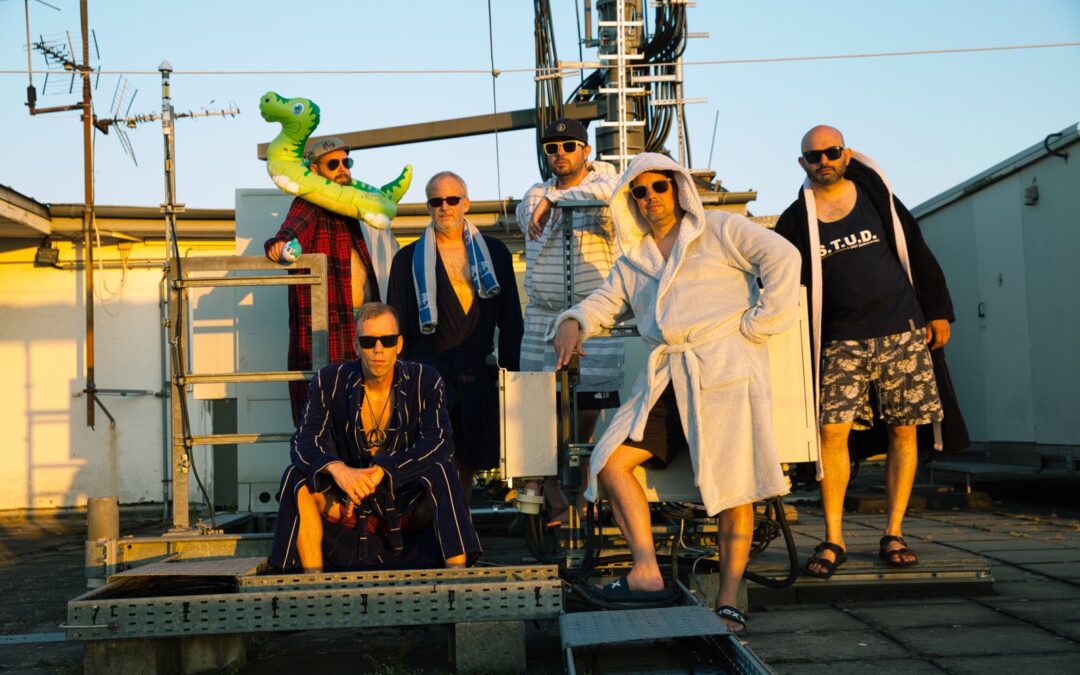 Group of men in robes and sunglasses posing on a rooftop with antennas and cables, one holding a green inflatable dinosaur