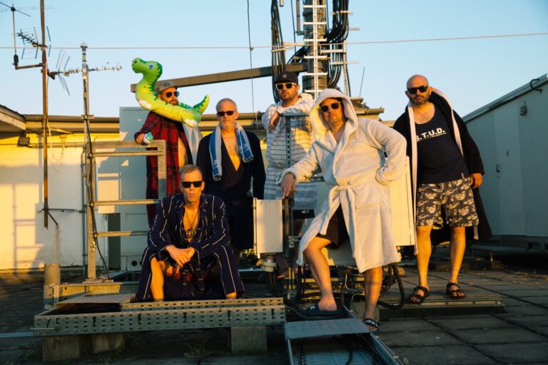 Group of men in robes and sunglasses posing on a rooftop with antennas and cables, one holding a green inflatable dinosaur