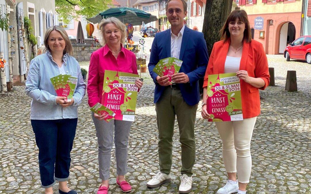 Four adults stand on a sunny cobblestone street in a European town, each holding colorful flyers for a Kunst Markt event and smiling at the camera.