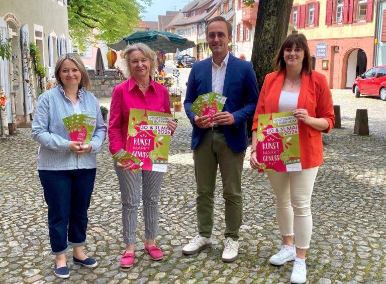 Four adults stand on a sunny cobblestone street in a European town, each holding colorful flyers for a Kunst Markt event and smiling at the camera.