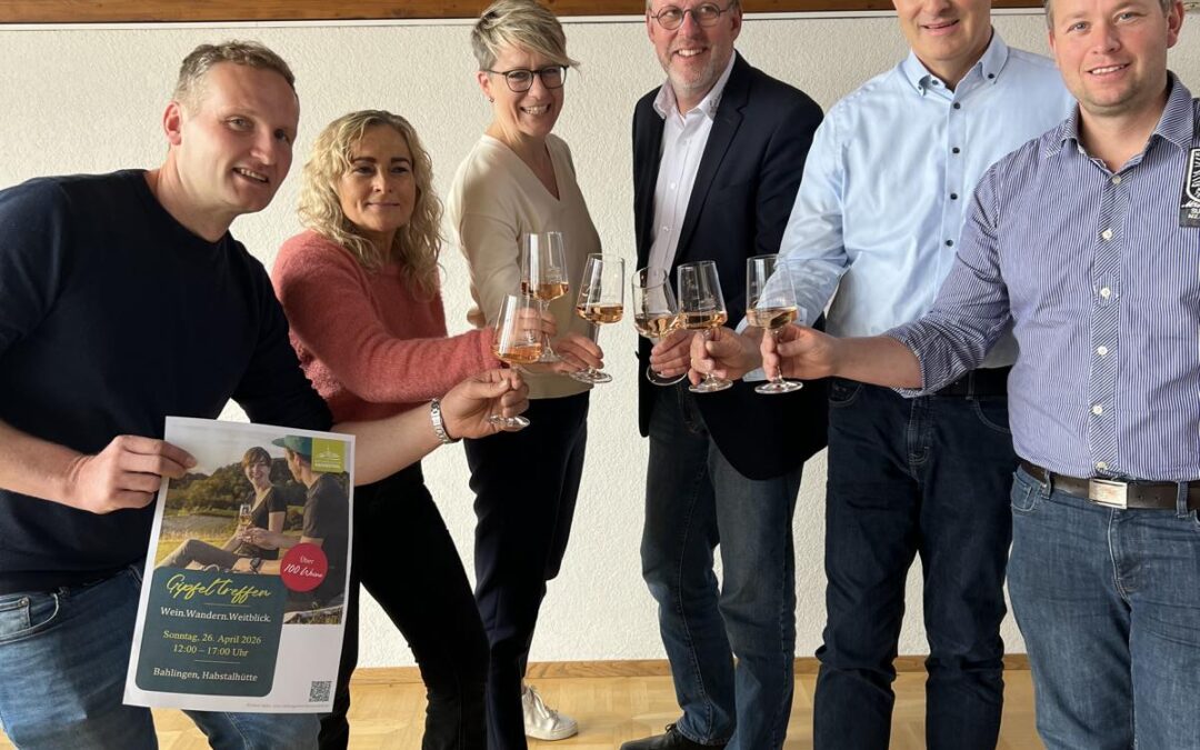 Six adults indoors raise wine glasses in a toast, with one person holding a promotional flyer/poster in front.