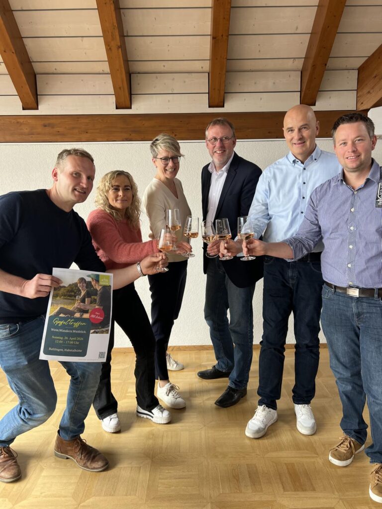Six adults indoors raise wine glasses in a toast, with one person holding a promotional flyer/poster in front.