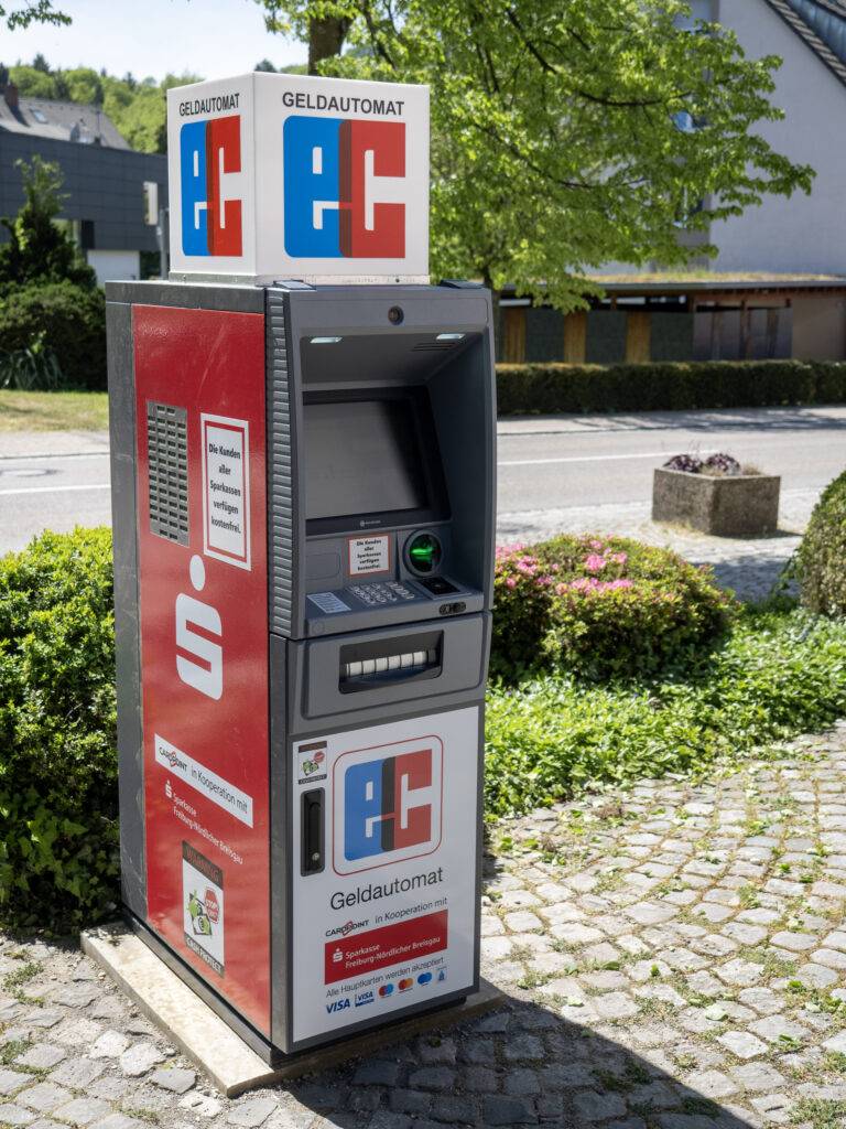 Outdoor cash machine (Geldautomat) in a freestanding vending kiosk, red side panel and blue/red logo, with greenery in the background.