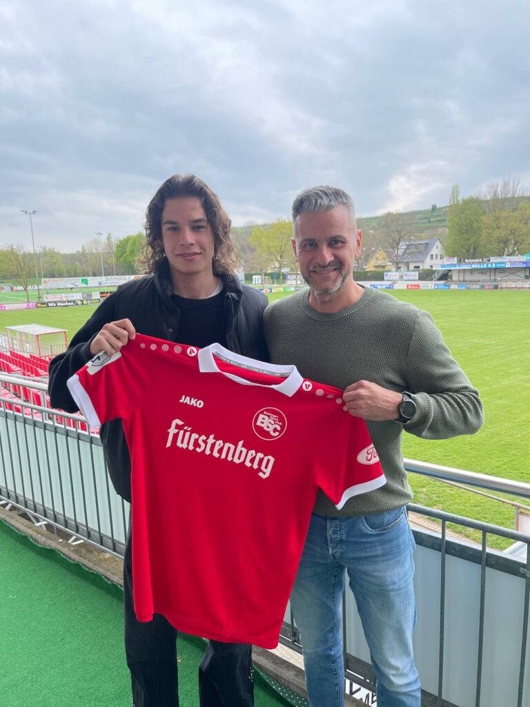 Two men stand on a stadium balcony smiling while holding a red soccer jersey with a white collar and Fürstenberg logo in the foreground.