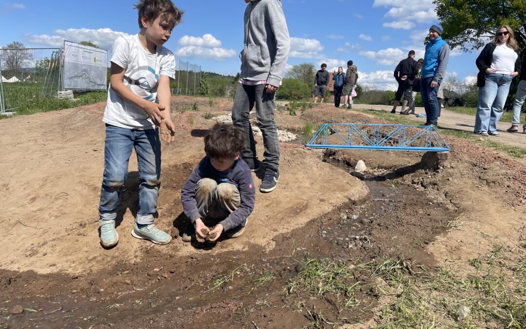 Children digging in a muddy area by a shallow stream while adults observe in the background on a sunny day.