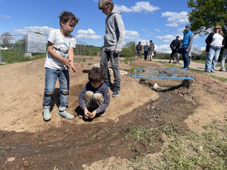 Children digging in a muddy area by a shallow stream while adults observe in the background on a sunny day.