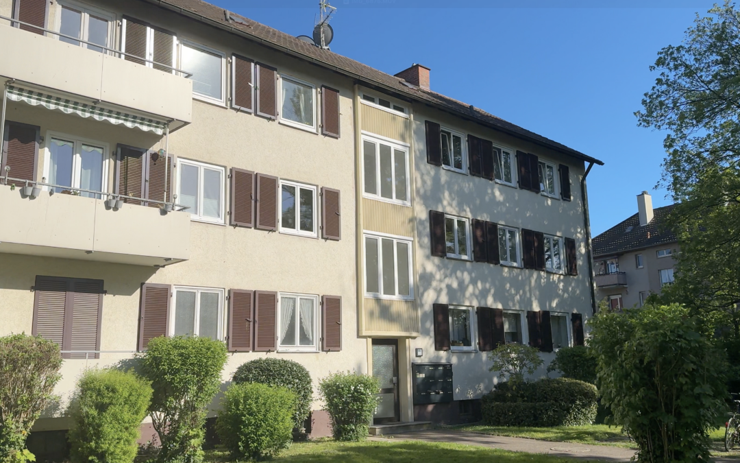 Residential apartment building with beige exterior, brown shutters, and multiple windows under a blue sky, with shrubs in front.